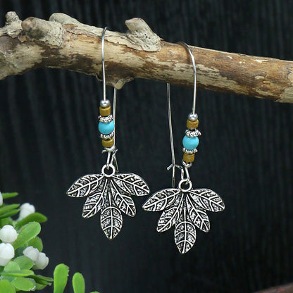 Silver leaf earrings with turquoise and brown beads hanging on a branch against a dark background.