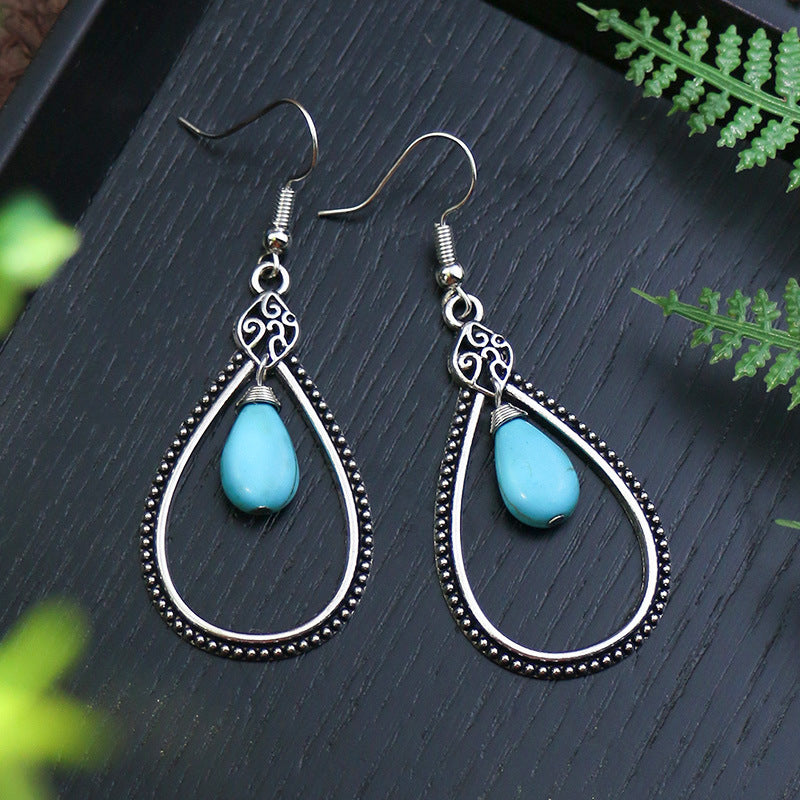 Teardrop-shaped earrings with turquoise stones on a dark surface with green leaves.